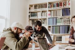 A group of young, male office employees in casual attire laugh and smile while working together on a project in a modern, bright collaboration space. Photo by Priscilla Du Preez via Unsplash.