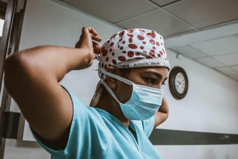 A Hispanic female healthcare worker secures the strings of her face mask behind her head. She is wearing pale blue scrubs and a head covering with a read and white pattern. Photo taken by SJ Objio via Unsplash.