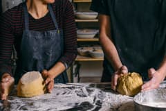 Two people knead dough on a well-floured surface in a commercial kitchen.