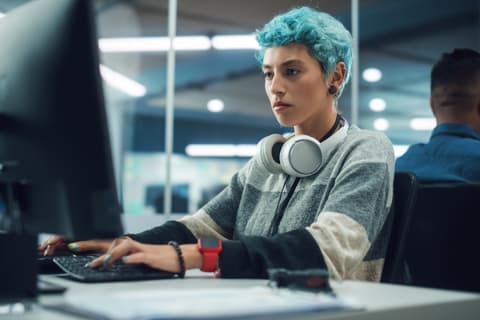 Portrait of young, feminine person with light blue hair focused on working at a computer in a modern office.