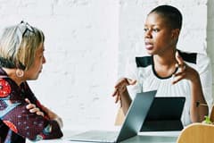 Image of two business women of color engaging in a discussion at a modern meeting table with a laptop on it.