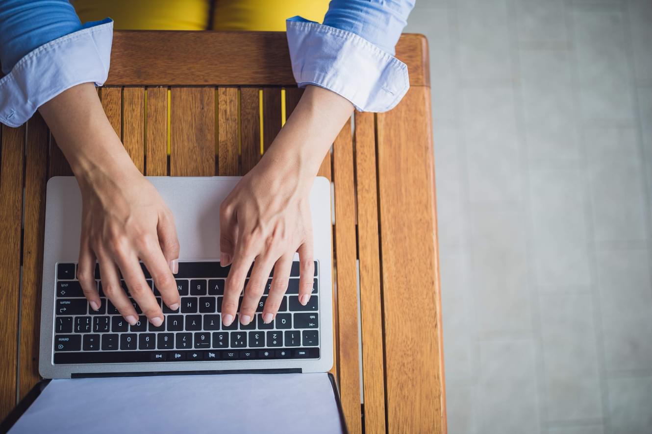 Overhead closeup of a person working on their computer, with their hands on top of the keyboard, and the computer is resting on a wooden table.