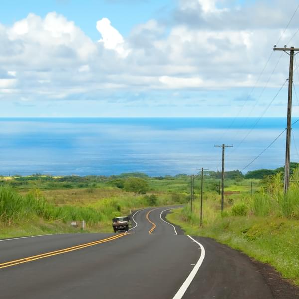 Empty road with power lines in Hawaiian countryside with car and ocean in background