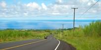 Empty road with power lines in Hawaiian countryside with car and ocean in background