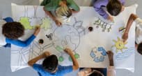 Small group of school aged children sit around a desk as they work together to color in a drawing.