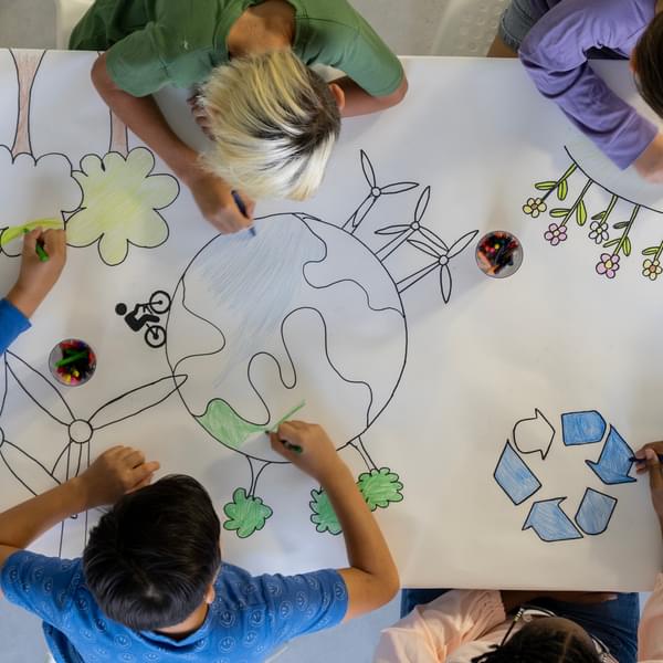 Small group of school aged children sit around a desk as they work together to color in a drawing.