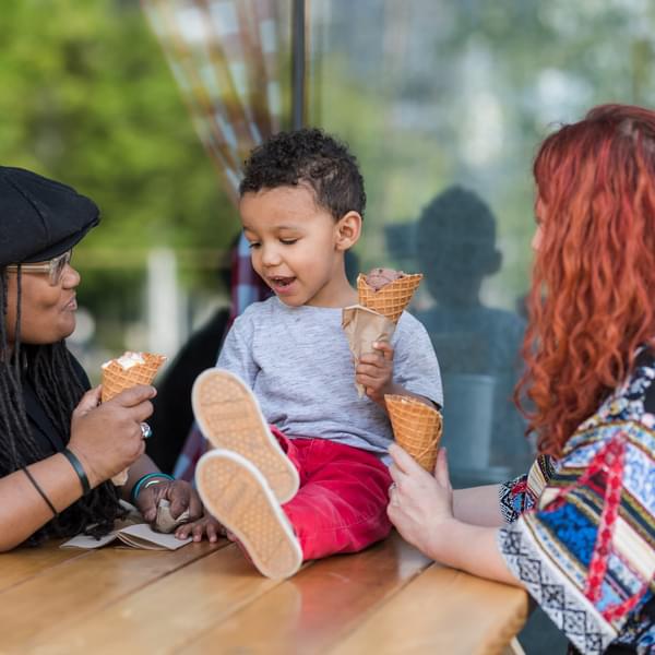 A homosexual biracial couple sit at a picnic table with their son and eat ice cream cones.