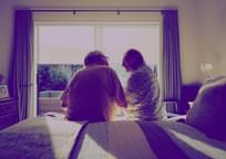 Back shot of senior couple sitting on edge of bed, in front of window