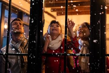 Child looking at the window and first snowflakes with family. Young kid girl sitting in living room at home, watching snow falling outside.