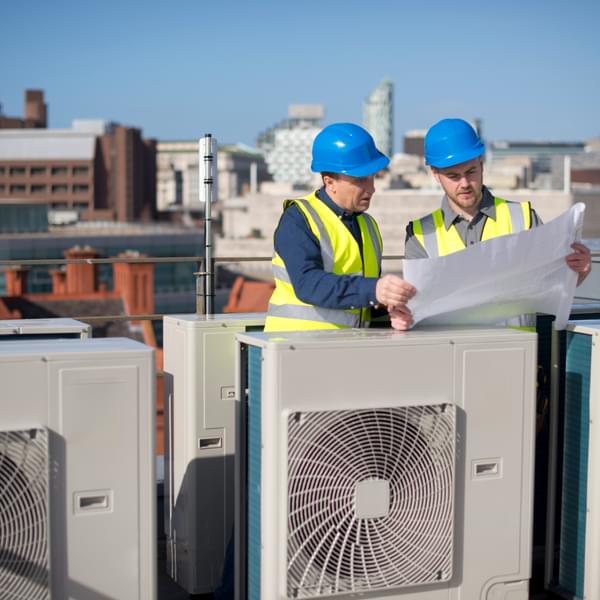 Two engineers on a rooftop next to HVAC equipment holding plans