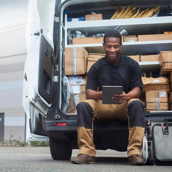 A service engineer sat at the back of his van using a digital tablet outside factory buildings