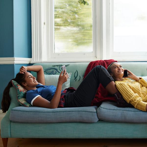Smiling women lounge on a sofa while using their phones.