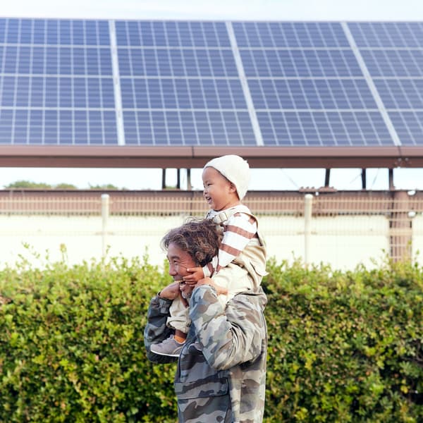 A senior man laughs as he carries a baby boy on his shoulders beside solar panels.