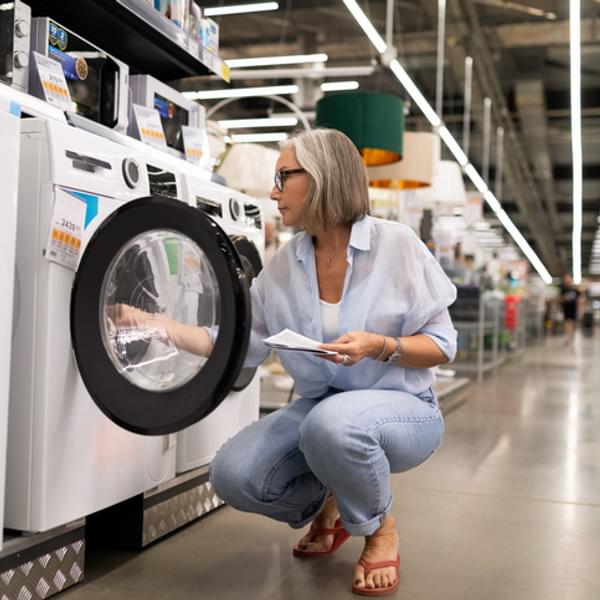 A woman squats by a washing machine in a well-lit appliance store, carefully inspecting the features while holding a product brochure. Bright overhead lights illuminate the space.