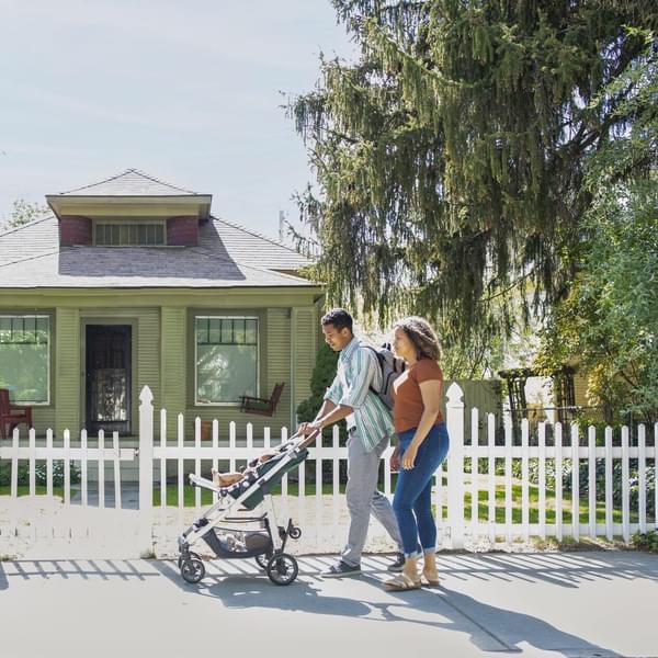 Young mother and father walking with baby daughter in stroller on neighborhood sidewalk.