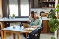 A mature man is working on the laptop and his wife is next to him while they are looking at a laptop