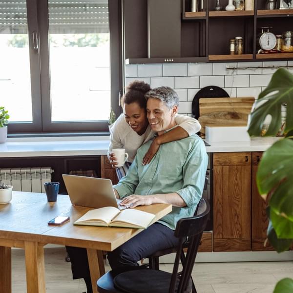 A mature man is working on the laptop and his wife is next to him while they are looking at a laptop