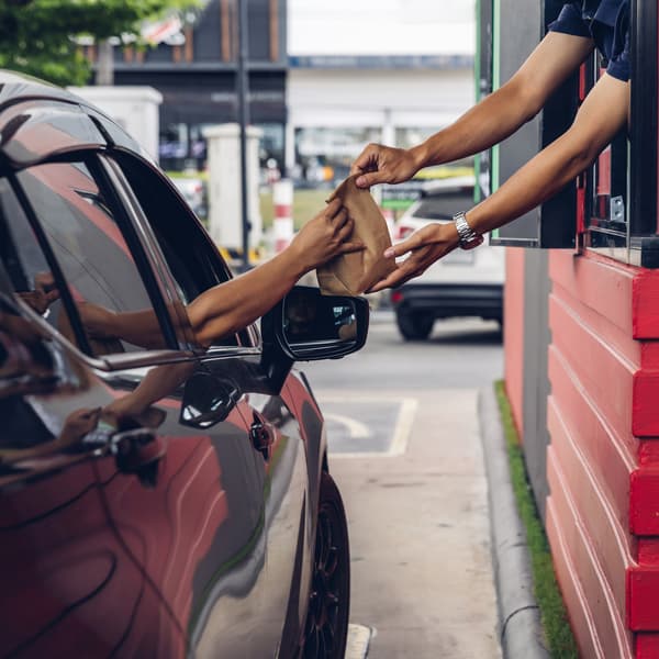 Employee handing a man in a car a bag of food through a fast food restaurant window.