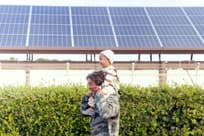 A senior man laughs as he carries a baby boy on his shoulders beside solar panels.