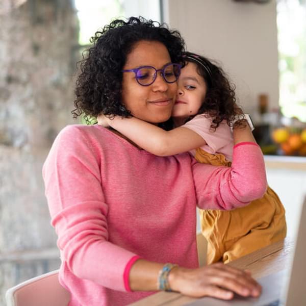 woman sitting at a table typing on computer while her toddler daughter climbs on her lap