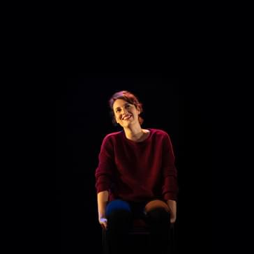 Phoebe Waller Bridge sits on a stool on a dark stage, smiling at the camera