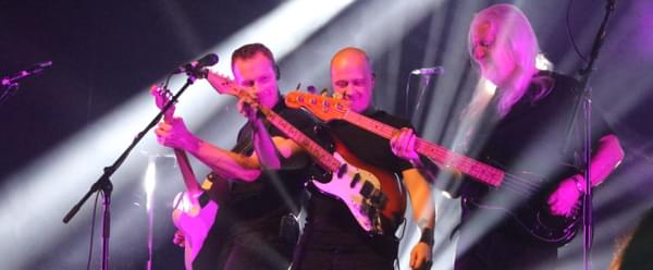 Three middle aged men play guitars on stage