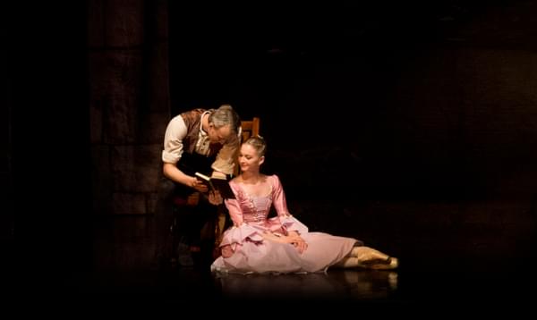 A male dancer sitting on a wooden chair holding a book. There is a female dancer in a pink dress is sat on the floor next to him and they are both looking at the book smiling
