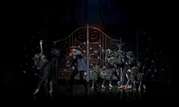 A group of dancers some in wolf costumes surrounding a scared male dancer who is trying to open a gate