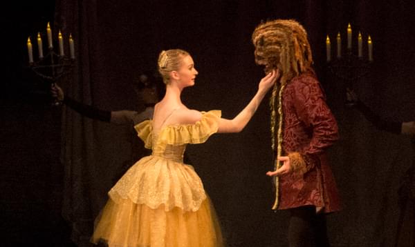 A female ballet dancer in a yellow dress touching the face of a male dancer in a beast costume