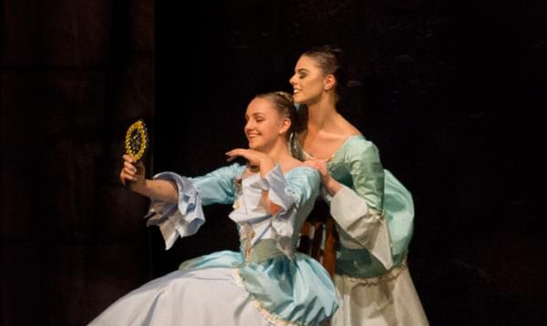 Two ballet dancers in white and blue dresses looking into a handheld mirror smiling