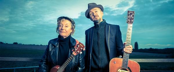 A man and a woman stand outdoors, side by side, holding a ukulele and a guitar