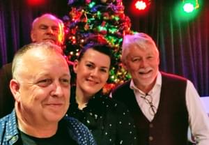 The four band members pose, smiling, in front of a Christmas tree