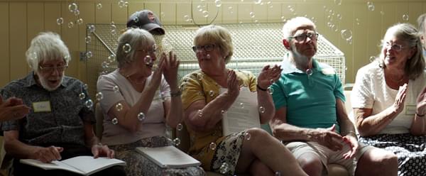 A group of older people sit, clapping, in front of floating bubbles