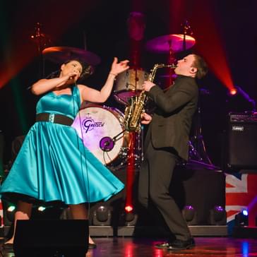 A woman in a bright dress sings on stage next to a man playing saxophone