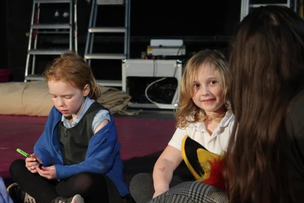 Two young girls sit cross legged on the floor. One holds a pen, concentrating; the other smiles at somebody just out of shot