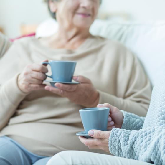 Two women sit chatting, each holding a cup of coffee, on a comfortable sofa