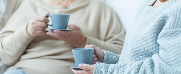 Two women sit chatting, each holding a cup of coffee, on a comfortable sofa