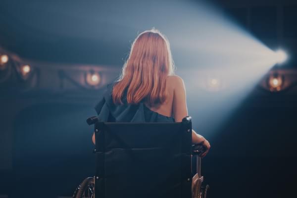 A woman in a wheelchair sits, back to the camera, on a stage in front of a spotlight