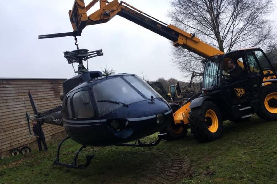 A blue helicopter without rotor blades being lifted by a JCB crane