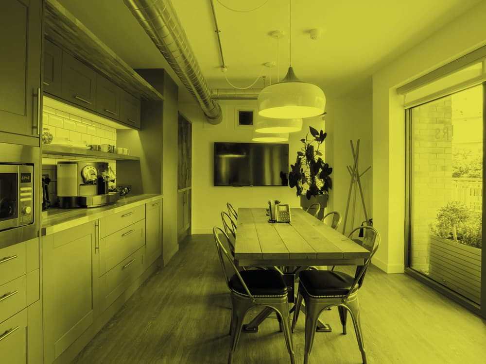 Interior view of a kitchen area. A long galley table sits centrally lit by multiple overhead handing lights. A kitchen covers the entire length of the room (on the left) with full height windows on the right. The image has a yellow filter applied.