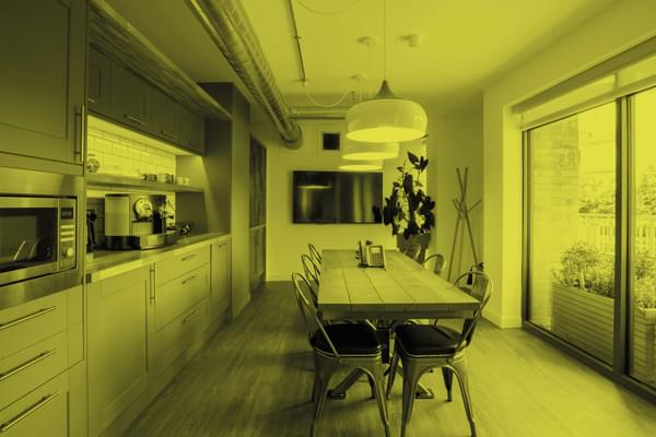 Interior view of a kitchen area. A long galley table sits centrally lit by multiple overhead handing lights. A kitchen covers the entire length of the room (on the left) with full height windows on the right. The image has a yellow filter applied.