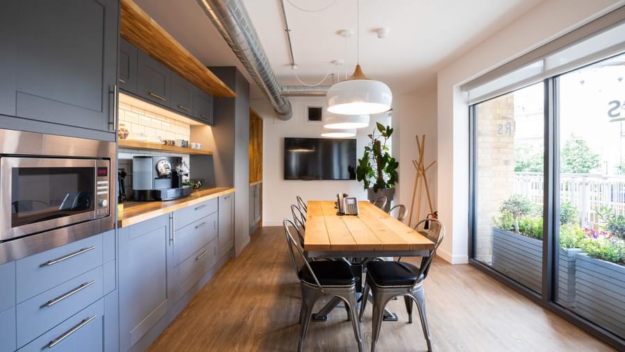 Modern kitchen with grey cupboards a wooden floor, wooden worktops and a large wooden table centred under three hanging ceiling lights.