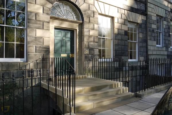 A grand stone-clad townhouse with a green door and a three-step stone staircase leading up to the door from the pavement. The front of the house is separated from the pavement by a black iron fence.