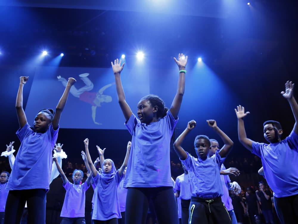 Many children singing with their arms up on a blue-lit stage. An illustration of a man breakdancing is the backdrop.
