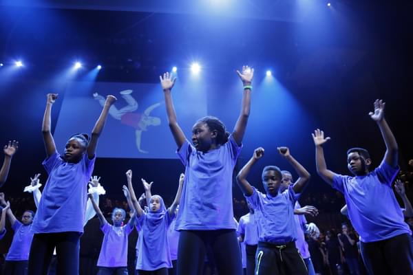Many children singing with their arms up on a blue-lit stage. An illustration of a man breakdancing is the backdrop.