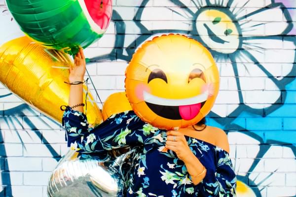 Woman holding smiling emoji balloon over her face.