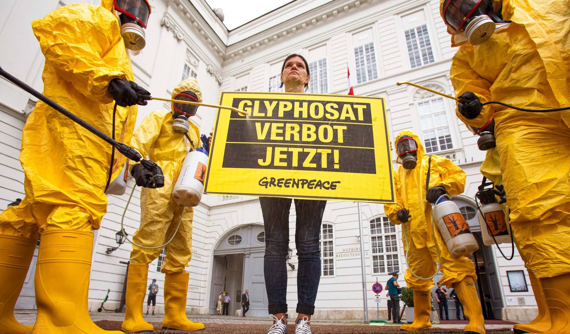 Menschen in gelben Anzügen und Gummistiefeln demonstrieren mit Schutzmasken und Spritzgeräten gegen Glyphosat
