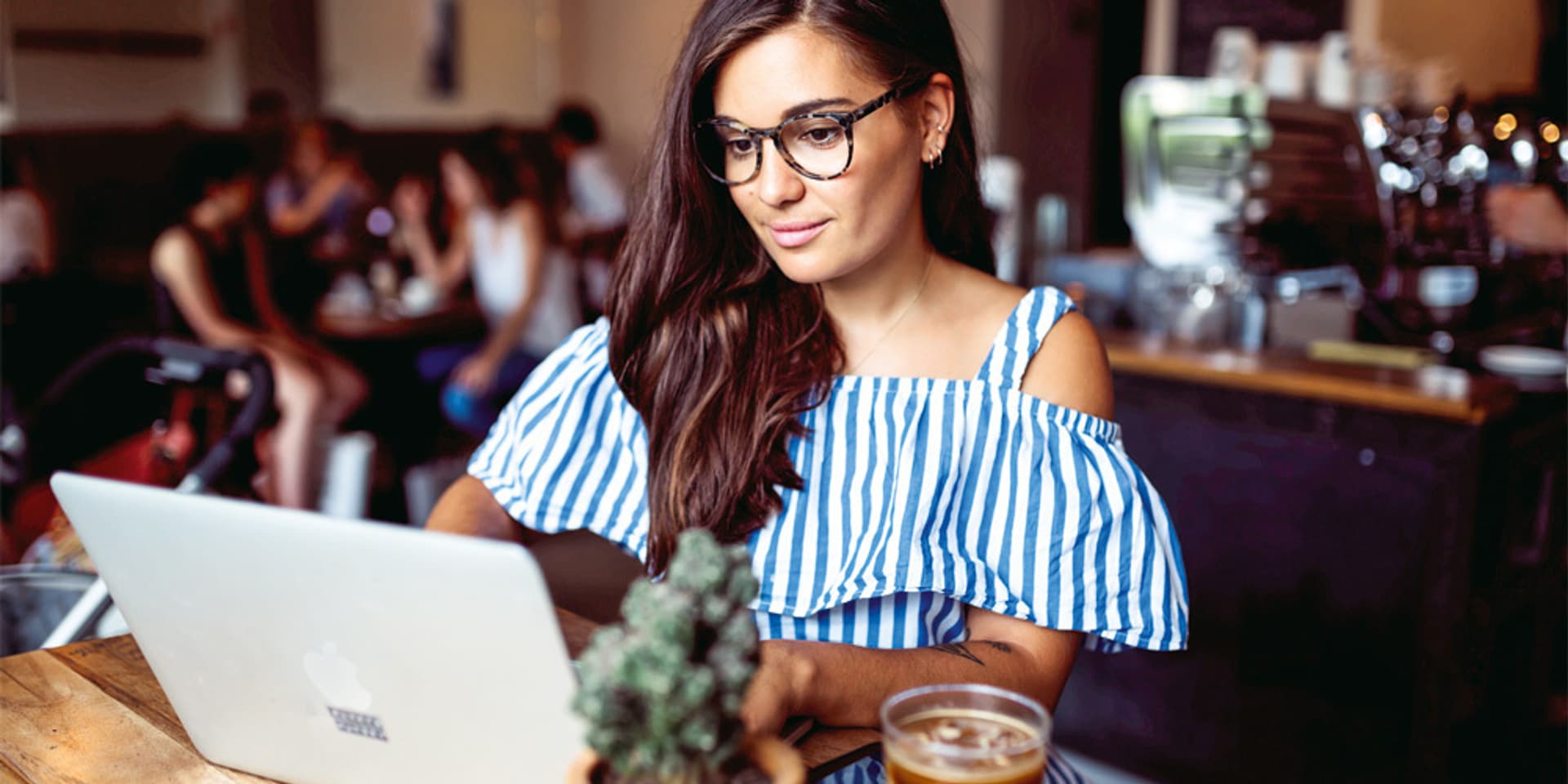 Eine Frau mit langem braunen Haar und dunkler Brille sitzt an ihrem Laptop in einem Café
