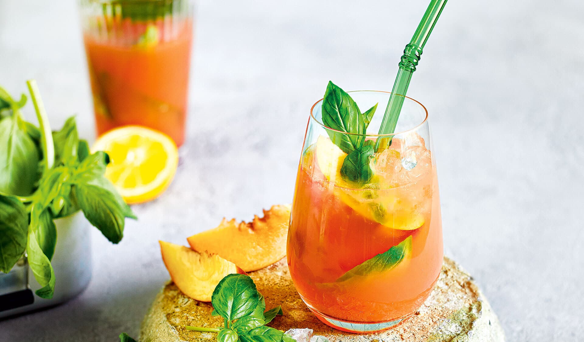 Glas mit orangenfarbenem Cocktail, garniert mit Basilikum, mit grünem Trinkhalm