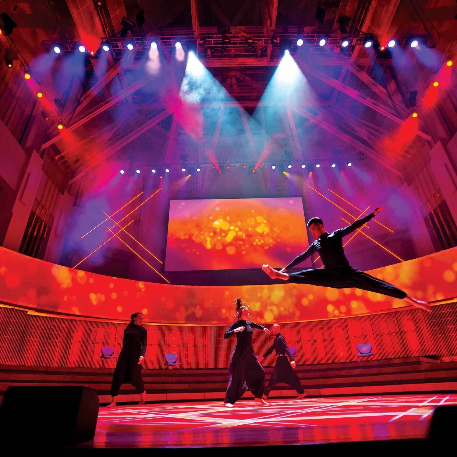A dancer leaps through the air in front of other performers on a stage lit with shades of orange and red